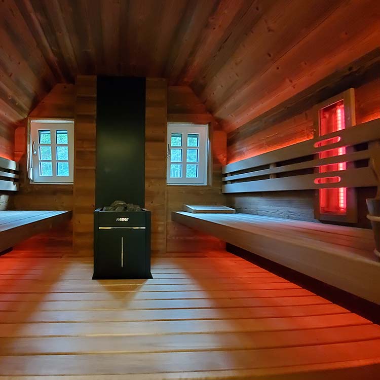 Innenraum der Holzsauna mit Bänken, Heizgerät, roter Akzentbeleuchtung und zwei kleinen Fenstern.