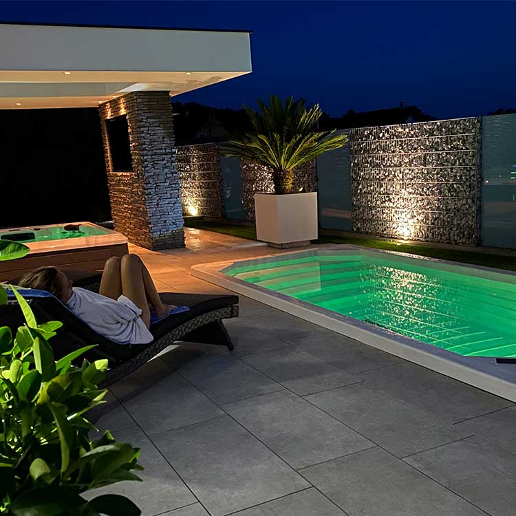 Frau, die sich nachts auf einem Liegestuhl am beleuchteten Swimmingpool entspannt, mit moderner Außendekoration.