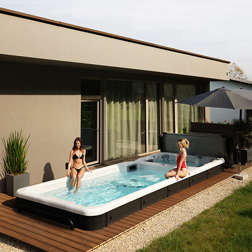 Swim Spa mit zwei Zonen, im Boden versenkt, auf einer Terrasse vor der Villa. Zwei Junge Damen sitzen am Beckenrand des Pools und genießen die Sonne.