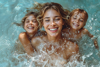 Eine Mutter hat viel Spaß mit ihren beiden Kindern im Swimmingpool. Sie planschen im Wasser und strahlen über das ganze Gesicht.