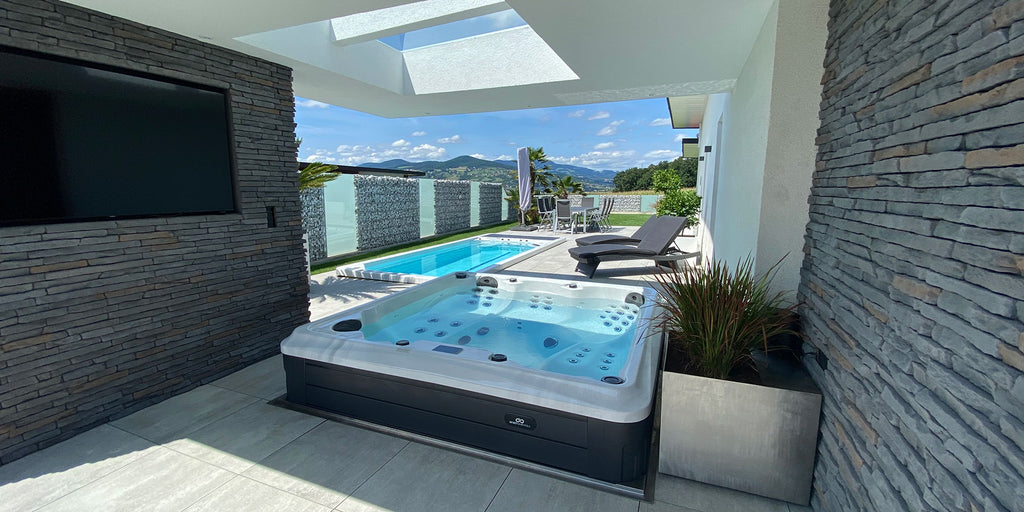 Moderne Außenterrasse mit Whirlpool, Pool, Liegestühlen, TV und Bergblick unter einem Dachfenster.