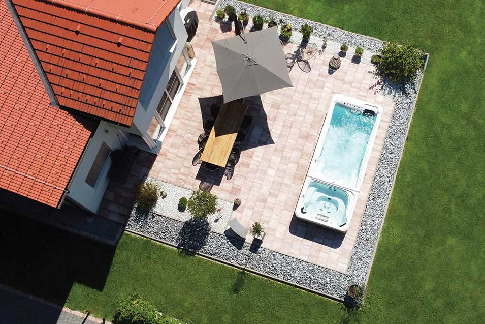 Artesian Swim Spa und Whirlpool Kombination auf einer gepflasterten Terrasse neben einem Haus mit rotem Ziegeldach, umgeben von grünem Rasen.