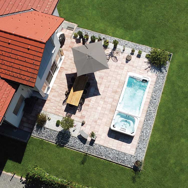Artesian Swim Spa und Whirlpool Kombination auf einer gepflasterten Terrasse neben einem Haus mit rotem Ziegeldach, umgeben von grünem Rasen.