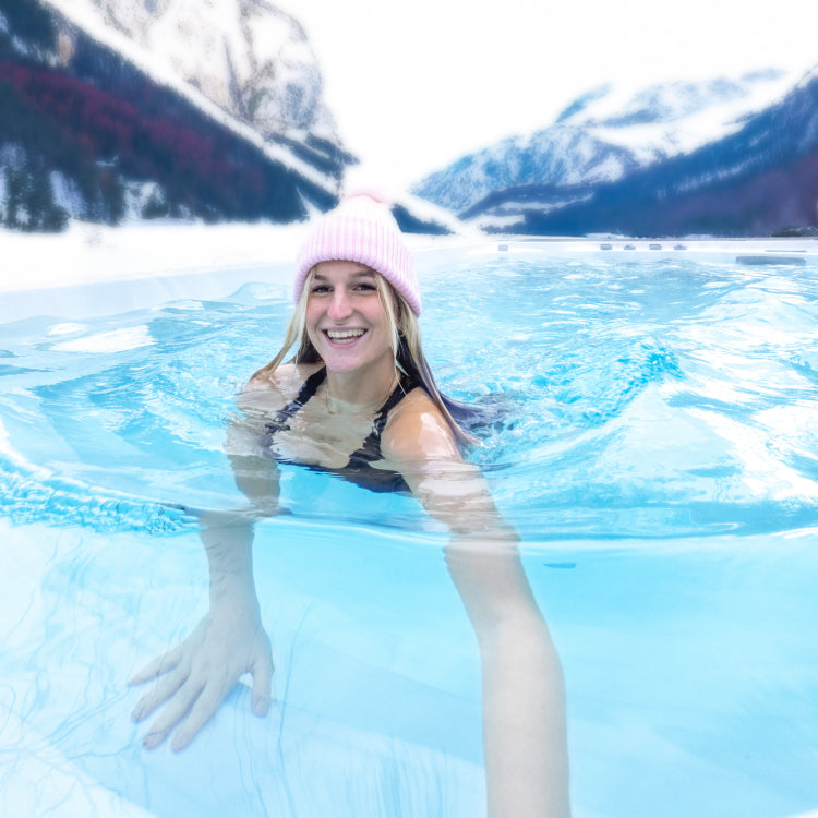Eine Frau mit rosa Mütze schwimmt lachend in ihrem Swimmingpool an einem kalten Wintertag in den Alpen.