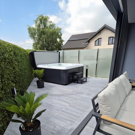 Outdoor-Whirlpool freistehend auf einer Terrasse mit Blick auf ein Wohnhaus und Garten, umgeben von Glasgeländer und Pflanzen.