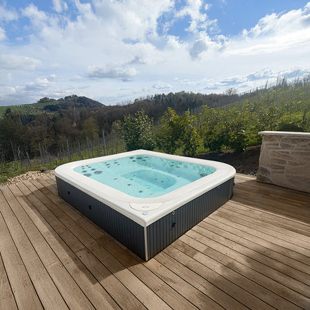Halb eingelassener Whirlpool auf einer Holzterrasse mit Aussicht über hügelige Landschaft und Wald im Hintergrund.